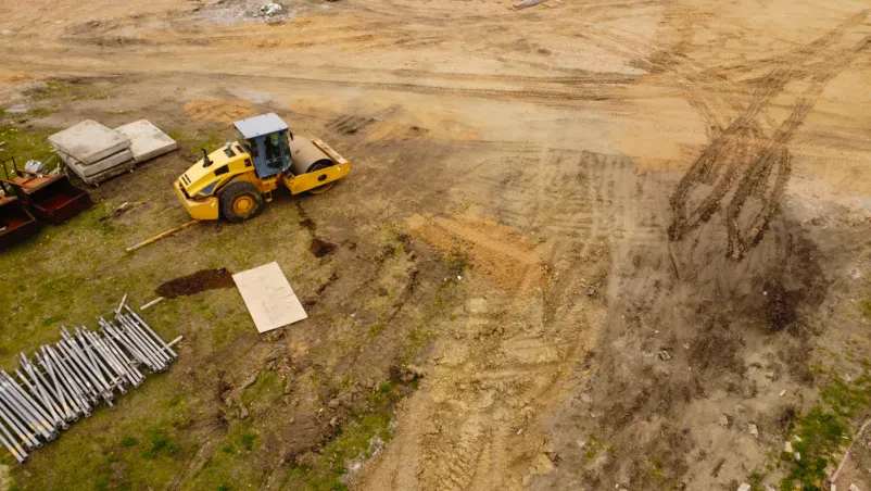 Pogled iz zraka na valjar na gradbišču Ptičja perspektiva prikazuje rumen valjar, ki utrjuje zemljo na gradbišču, obdanem z različnimi gradbenimi materiali. Delavci skrbno urejajo površino in pripravljajo teren za nadaljnja dela.