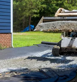 Traktor raztresa gramoz na črno ponjavo ob novozgrajeni hiši. To je pomemben del priprave terena za asfaltiranje Štajerska, saj se s tem zagotavlja pravilna drenaža in utrjenost podlage.