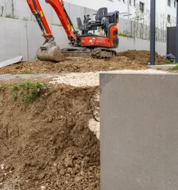 Mini bager skrbno pripravlja in ureja zemeljsko površino v bližini moderne stanovanjske stavbe. Opravlja manjše gradbene izkope in ravnanje terena za urejeno okolico, kot je razvidno iz slike.