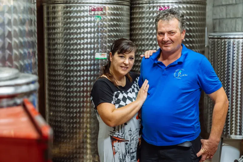 Portret nasmejanega para, Melinde in Denisa Jurada, ki ponosno stojita ob svojih vinogradih. Predstavljata mlajšo generacijo vinogradnikov in pridelovalcev olja.