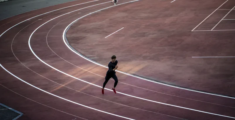 Prizor od zgoraj prikazuje atleta med šprintom na rdeči atletski stezi. Poudarjena sta hitrost in dinamika gibanja med treningom.