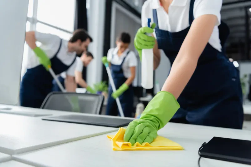 Čiščenje pisarniških površin s čistilom Čistilka z rokavico v roki drži stekleničko detergenta in krpo, pripravljena za temeljito čiščenje mize v sodobni pisarni.