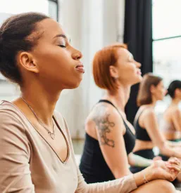 Mlada ženska z zaprtimi očmi meditira ob prijateljicah v jogijskem razredu, ki se sproščajo v enostavnem položaju, poudarjajoč ravnovesje in vitalnost.