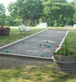 Prostrano zunanje balinarsko igrišče s peščeno podlago, obdano z zelenjem in praznimi stoli, čaka na igralce in gledalce v prijetnem ambientu.