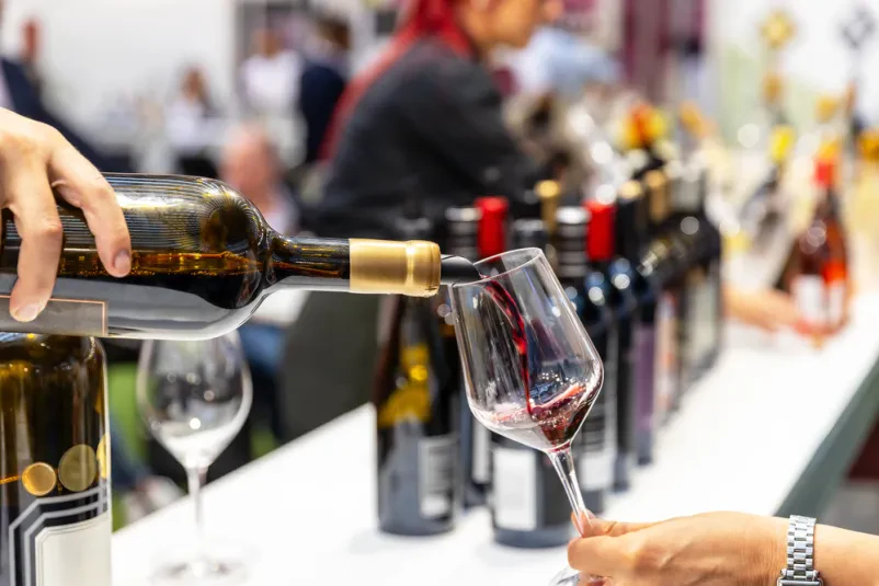 Sommelier toči rdeče vino gostom Uslužbenec, sommelier, s profesionalno natančnostjo toči rdeče vino v kozarce obiskovalcem na dogodku. Gostje v ozadju že okušajo vino, kar kaže na živahno degustacijo vin Turnišče, Prekmurje.