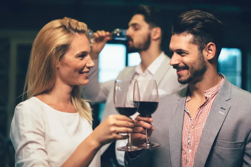 Skupina ljudi na pokušini vin Nasmejana skupina prijateljev uživa v sproščeni degustaciji vin v vinarni. Vsak udeleženec drži kozarec in se pogovarja, kar poudarja družabni vidik dogodka, ki je pogosto del promocije vinogradništva.