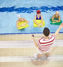 Skupina otrok s plavalnimi pripomočki v vodi pozorno posluša navodila svojega trenerja. Skupinska vadba spodbuja socialne veščine in zabavo.