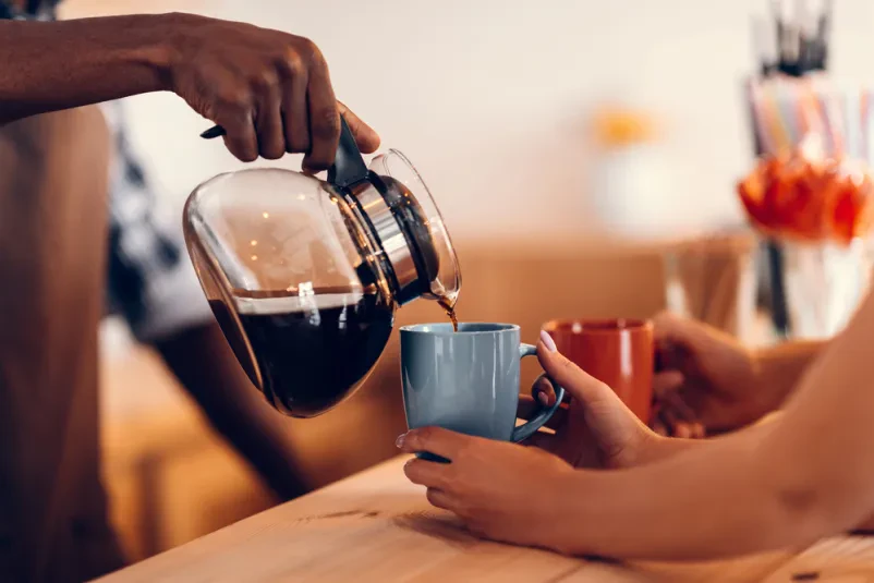 Prijazen barista s poudarkom na natančnosti naliva dišečo kavo v elegantne skodelice, kar je značilno za Dobro okrepčevalnico Maribor, Štajerska, kjer strežba pijače poteka s srcem.