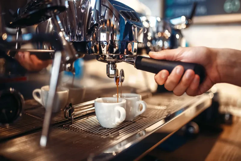 Barista s spretnimi rokami pripravlja osvežilno kavo z uporabo profesionalnega kavnega avtomata na svetlem pultu lokala. To je slika jutranje svežine in priprave toplih napitkov.