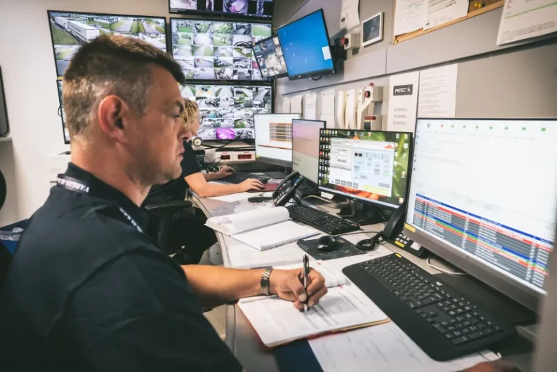 Dva uniformirana varnostnika usklajujeta dejavnosti prek radijskih postaj, kar zagotavlja učinkovito in neprekinjeno komunikacijo. Njihovo budno varovanje je ključnega pomena.