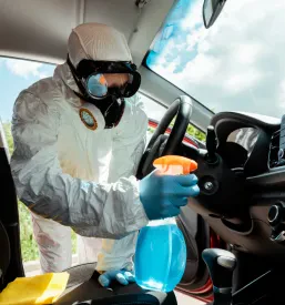 Specialist v zaščitni obleki z razkužilnim sprejem in krpo natančno čisti notranjost avtomobila, zagotavljajoč vrhunsko higieno in zaščito.