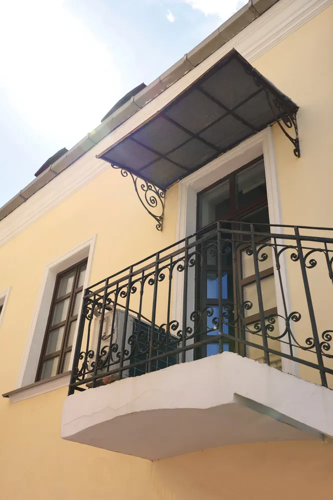 Kovana ograja balkona z nadstreškom Eleganten kovan balkon z zaščitnim nadstreškom in ograjo na rumeni fasadi. Odlični Kovinski nadstreški, ograje, stopnice Prekmurje za domove.