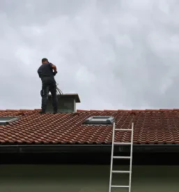 Samoten dimnikar na strehi hiše, ki skrbno izvaja čiščenje dimnika z dolgo krtačo pod jasnim nebom.