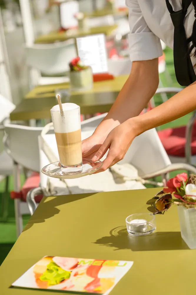 Natakarica s skrbnostjo postreže sveže pripravljeno kavo latte na leseni mizi zunanji terasi, poudarjajoč kakovost pijač.