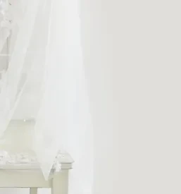 Prefinjena bela poročna obleka z dolgim pajčolanom nežno visi na stolu, pripravljena za pomemben dan. Poudarek je na detajlih in eleganci.