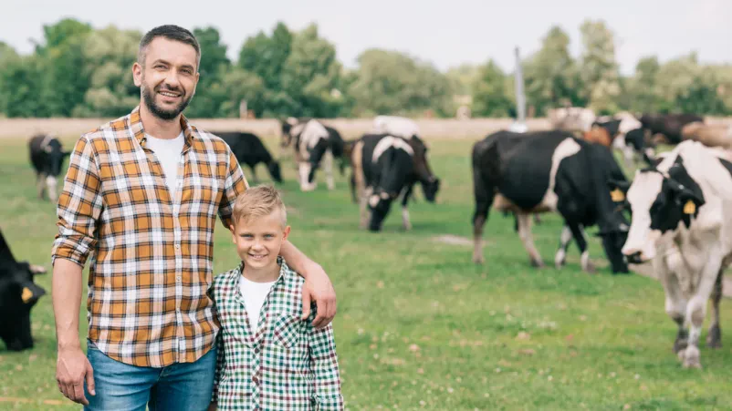 Nasmejan oče in sin ob govedu na kmetiji Srečen oče in sin se smehljata ob pašnih kravah, kar ponazarja Povezovanje in izobraževanje govedorejcev Sevnica, Posavje med generacijami.