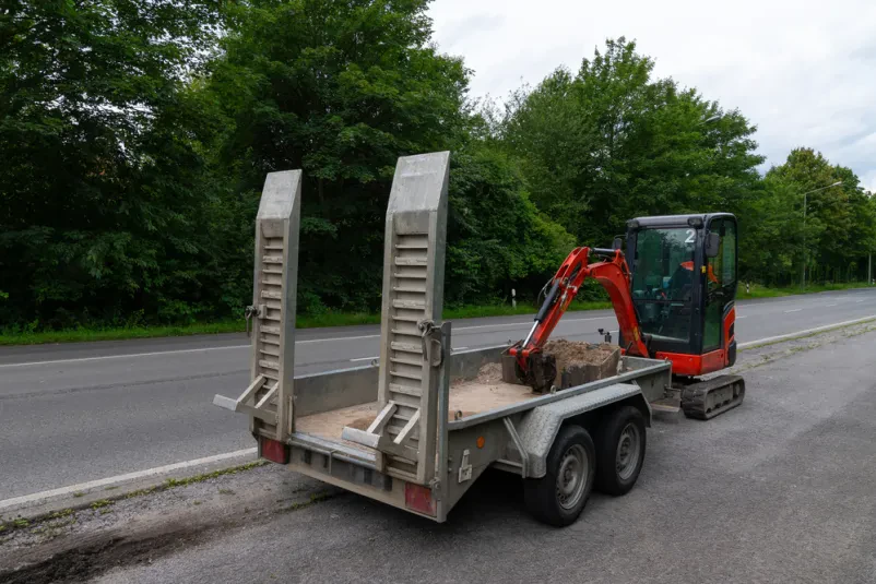 Priprava rdečega bagra za cestni transport Rdeč bager je naložen na prikolico ob cesti, pripravljen za prevoz. Rampi sta spuščeni, žlica bagra pa se dotika tal.