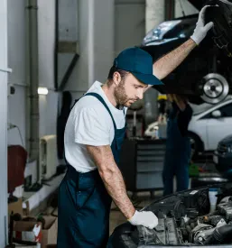 Avtomehanik v zaščitnih rokavicah podrobno pregleduje motor avtomobila, kar zagotavlja učinkovito popravilo vozil.