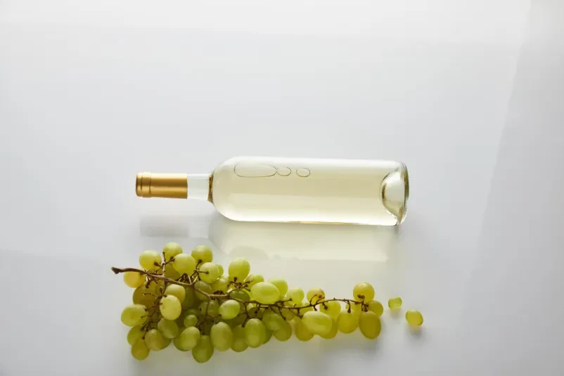 Steklenica belega vina z grozdjem Pogled od zgoraj na steklenico kakovostnega belega vina, postavljeno ob svežem grozdju na čistem belem ozadju. Poudarja svežino in kakovost pijače.