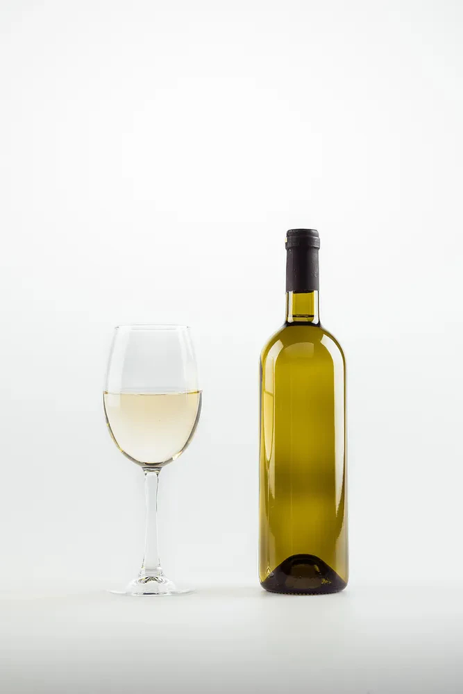Belo vino v kozarcu in steklenici Bližnji posnetek kozarca in steklenice belega vina, elegantno postavljenih na belem ozadju. Prikazuje preprostost in eleganco ob vsaki priložnosti.