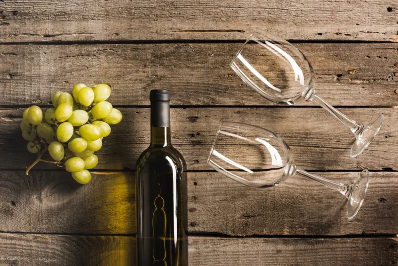 Belo vino, kozarci in grozdje na mizi Pogled od zgoraj na elegantno postavljeno mizo, kjer sta steklenica belega vina in dva kozarca obdana s svežim grozdjem in vintage prtom. Idealno za gurmanske užitke.