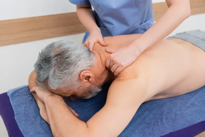 Masaža hrbta v rehabilitacijskem centru Izkušena maserka izvaja masažo hrbta srednjeveškemu moškemu v rehabilitacijskem centru, s poudarkom na odpravi bolečin in izboljšanju gibljivosti.