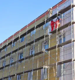 Gradbeni delavci na odru popravljajo fasadno toplotno izolacijo na zunanji steni stanovanjske stavbe. Takšna energetska obnova je ključna za zmanjšanje stroškov ogrevanja in izboljšanje učinkovitosti, kar ponuja ugoden fasader Obala.