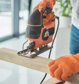 Približen posnetek moškega, ki s spretnostjo uporablja električno vbodno žago za obdelavo lesa, kar je pomemben del zaključnih gradbenih del pri prenovi.