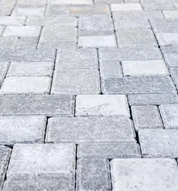 Utrjena površina pred objektom z rdečo fasado, kjer delavec, ki nosi zaščitno čelado, polaga tlakovce na tla.