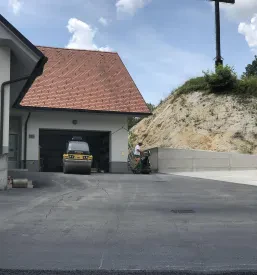 Asfaltirano dvorišče pred garažnim objektom stanovanjske hiše. Ob hiši je kup peska.