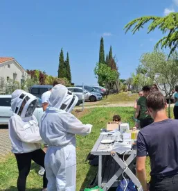 Mladi v čebelarski opremi stojijo okrog mize, kjer je predstavljena čebelarska oprema.