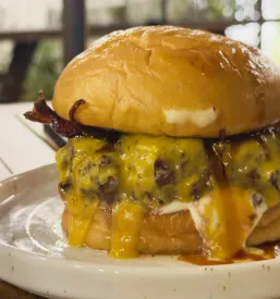 Okusen slaninast cheeseburger z obilico stopljenega sira na popečeni brioš štručki, ki vabi k uživanju v bogatem okusu.