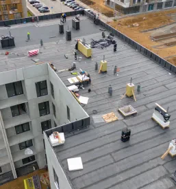 Posnetek z dronom prikazuje gradbišče z delavci, ki so zaposleni z deli na ravni strehi, kar ponazarja obsežnost projektov izdelave in montaže ravnih streh Ljubljana.