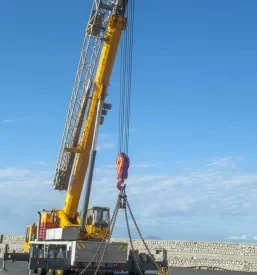 Močan rumeni mobilni žerjav na kolesih, ki stoji pod modrim nebom in čaka na dvig težkega tovora. Pripravljen je za zahtevna dela v industriji in gradbeništvu, s poudarkom na robustnosti in zmogljivosti.