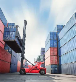 Velik ladijski kontejner, ki ga industrijski žerjav dviga na nakladalni pomol v prometnem pristanišču, kar je ključen del servisa industrijskih dvigal Slovenija za logistične potrebe.