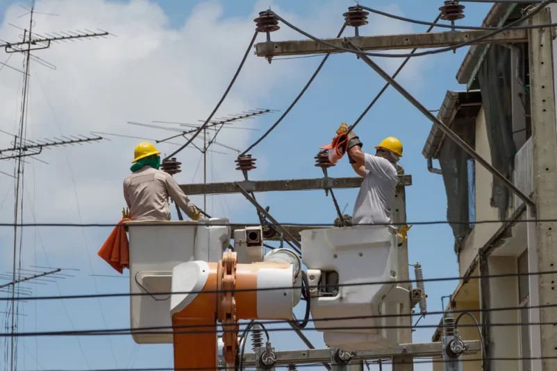 Električar v košari dvigala ob električnem drogu Izkušen električar varno namešča električne inštalacije na visokem stebru z uporabo košare dvigala, kar prikazuje kompleksnost servisa industrijskih elektroinštalacij in dvigal Slovenija.