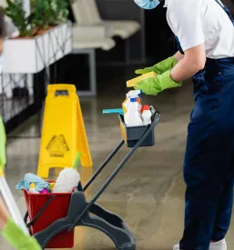 Čistilec z masko in rokavicami pripravlja čistila na vozičku v poslovnem prostoru.