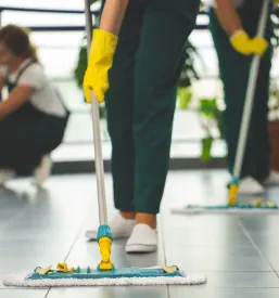Tri osebe v delovnih oblekah in rokavicah pomivajo tla v velikem prostoru.