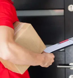 Moški dostavljavec v rdeči uniformi stoji pred vhodnimi vrati in drži velik rjav kartonski paket.