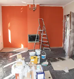 Pripravljena soba za pleskanje z oranžno steno, lestvijo in vedri barve.