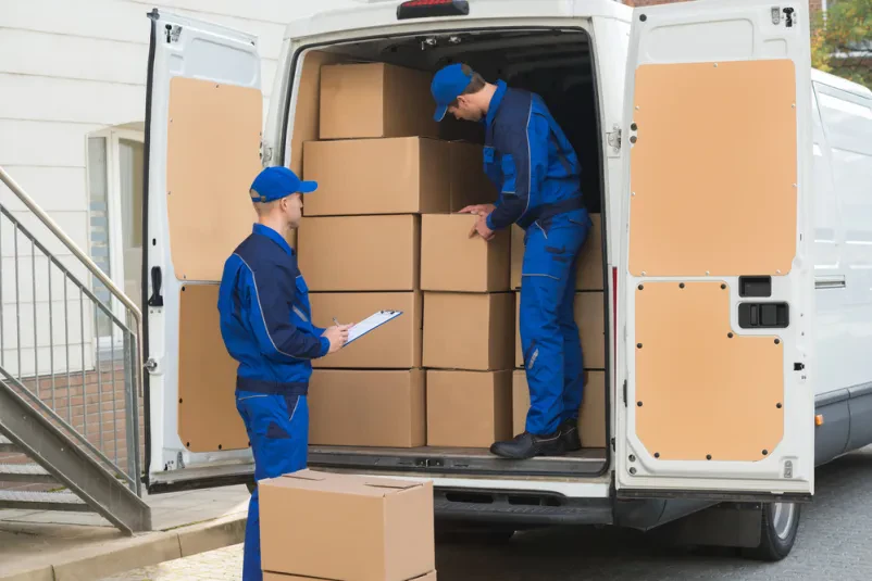 Dva dostavljavca v modrih uniformah marljivo razkladata kartonske škatle iz tovornega vozila. Njuno usklajeno delo je ključno za hiter transport in prevoz pošiljk po Sloveniji, ki zagotavlja pravočasno dostavo.