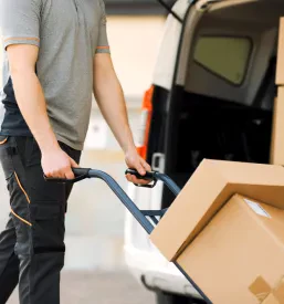 Profesionalni kurir v modri uniformi in s ščitnico na kapi s koncentracijo potiska ročni voziček, naložen z mnogimi kartonskimi paketi. To ponazarja zanesljiv in hiter transport in prevoz pošiljk po Sloveniji.