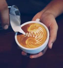Detajl skodelice kave z umetniškim latte artom, postavljene na leseno mizo v prijetnem ambientu, idealen za kavarno ali kava bar Gornja Radgona.
