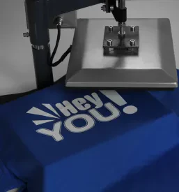 Podroben posnetek industrijske toplotne preše, ki je pripravljena za tisk logotipa ali grafike na modro majico, ležečo na čisti beli mizi, poudarja natančnost tiskarskega procesa.