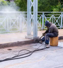 Delavec z zaščitno masko in opremo peska kovinsko konstrukcijo mostu. Ta postopek je ključen za vzdrževanje in podaljšanje življenjske dobe objekta.