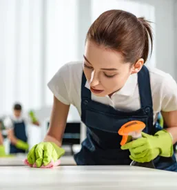 Ženska v predpasniku in rokavicah čisti pisarniško mizo s pršilom in krpo, v ozadju sta še dva čistilca.