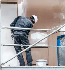 Delavec v beli čeladi in temnih oblačilih stoji na gradbenem odru in nanaša omet na fasado.