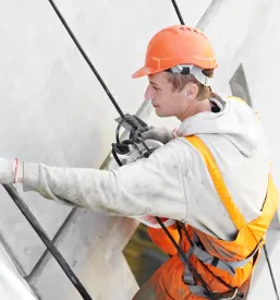 Delavec v oranžni zaščitni čeladi in varnostnem pasu se spušča ob steni stavbe.