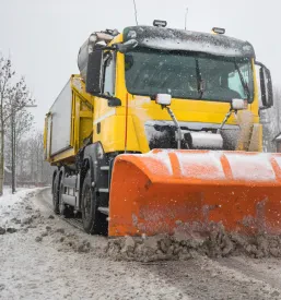 Rumeni tovornjak z oranžnim plugom čisti sneg z vaške ceste med sneženjem.